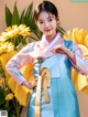 A woman in a blue and pink hanbok standing in front of yellow flowers.