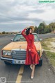 A woman in a red dress and straw hat leaning against a yellow car.